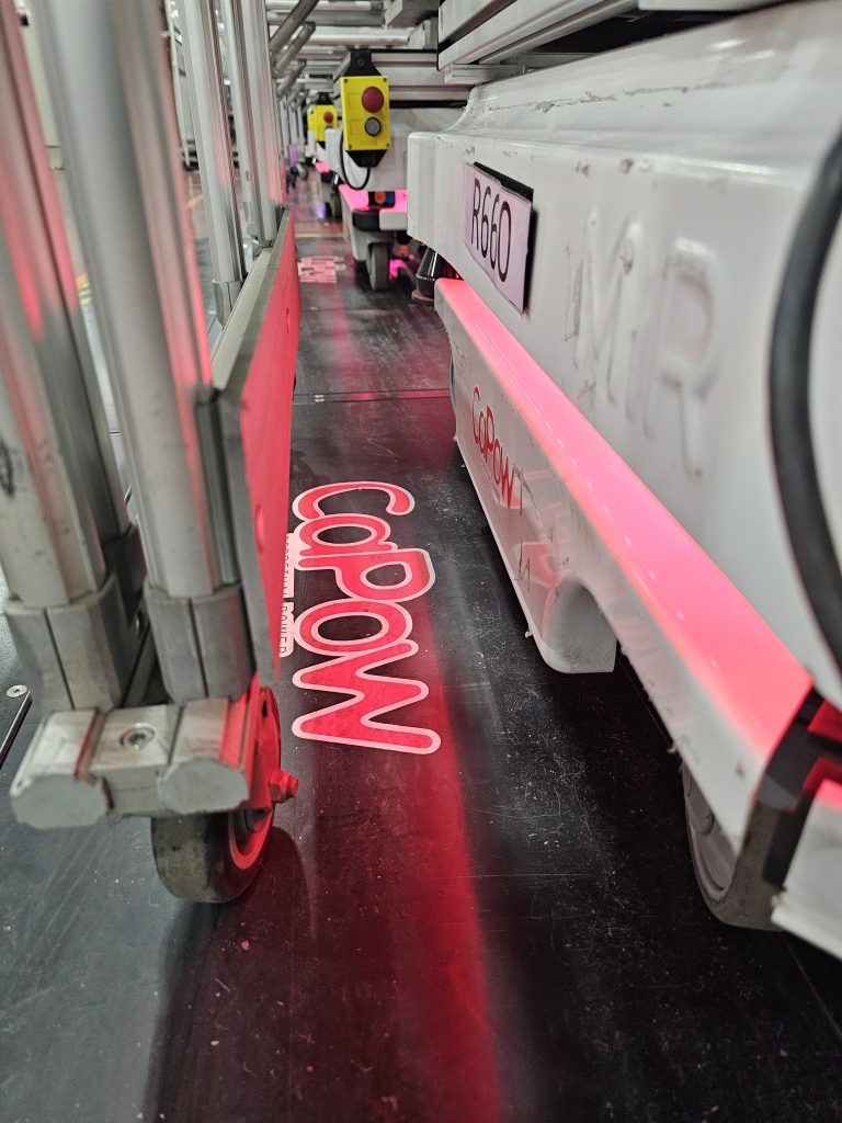 A close-up view of automated machinery with wheels, featuring a glowing logo on the floor and control buttons visible.