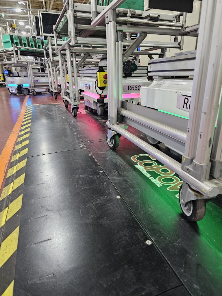 A row of automated carts with green and blue lights on a black floor, showcasing a warehouse or industrial setting.